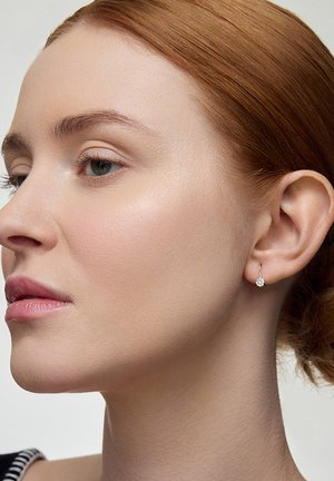 Srebrne kolczyki w kształcie kół z małym, okrągłym kamieniem szlachetnym. Gładka cera modelki i delikatny makijaż podkreślają biżuterię.