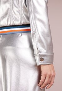 Silverfärgad metalljacka med en ribbad midja i blå, orange och vita ränder, samt en knappstängning på ärmen. Slät textur.
