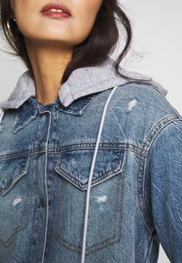 Veste en denim avec une capuche grise doublée. Les caractéristiques incluent des accents usés, deux poches poitrine et des détails de couture contrastants.