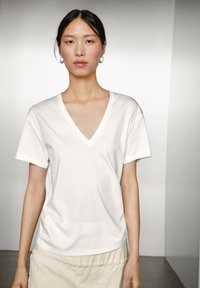Biała bawełniana koszulka typu V-neck z krótkimi rękawami, luźny krój i gładka textura, połączona z neutralną spódnicą z miękkiego materiału.