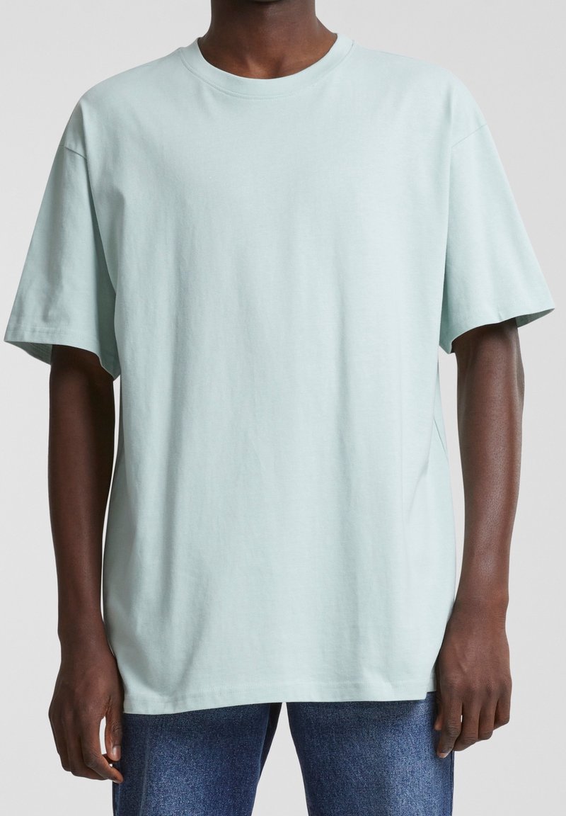 Homme portant un t-shirt uni bleu clair oversize à manches courtes avec un col rond et un jean bleu sur un fond uni.