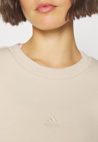 Femme portant un sweat à capuche beige Adidas avec le logo centré sur la poitrine, cheveux bruns courts et couleur de lèvres neutre.