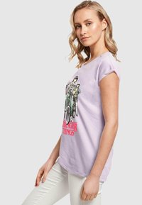 Lilac T-Shirt mit kurzen Ärmeln, featuring einen Grafikdruck von Charakteren auf Fahrrädern, mit "Stranger Things"-Schriftzug in fettem Pink. Weicher Stoff, lockere Passform.