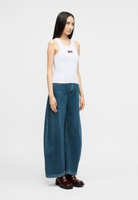 Γυναίκα που φοράει λευκή αμάνικη μπλούζα με μικρό κόκκινο κεντητό τίγρη, φαρδιά μπλε τζιν παντελόνια και σκούρα κόκκινα μποτάκια μέχρι τον αστράγαλο, στέκεται πλευρικά.