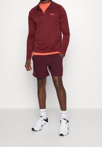 Maroon långärmad zip-up-tröja över en orange skjorta, matchad med maroon idrottsshorts och vita sneakers med svarta detaljer.