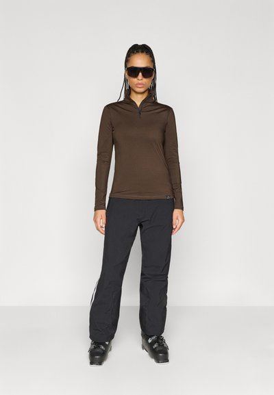 Καφέ μακρυμάνικη φούτερ με φερμουάρ συνδυασμένος με μαύρο παντελόνι που έχει λευκές πλευρικές ρίγες, και μαύρες μπότες σκι. Το μοντέλο φορά σκούρα γυαλιά ηλίου.