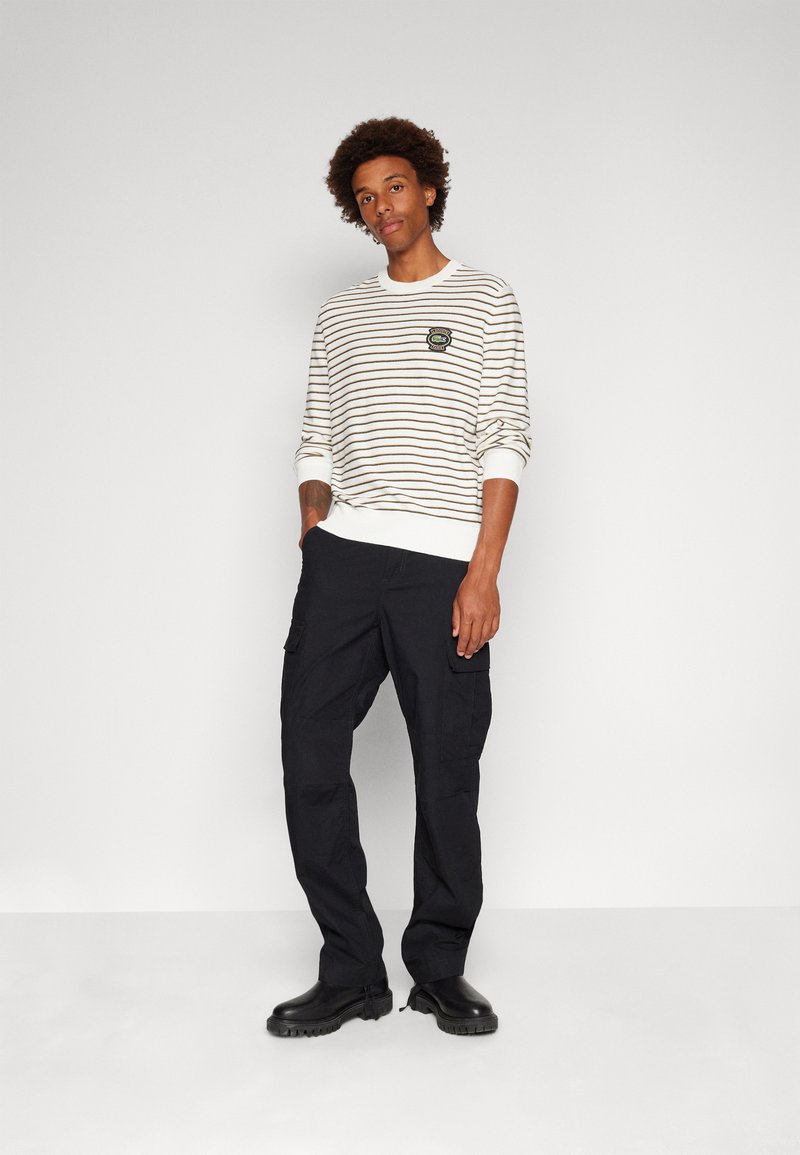 Gestreepte crewneck sweater in crème en bruin, met een geborduurd patch, gecombineerd met zwarte cargobroek en zwarte enkellaarzen.