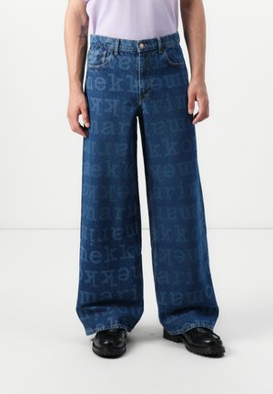 Wide-leg blue jeans made from denim, featuring a repeated logo pattern, five pockets, and a button fly, paired with black shoes.