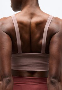 Μυώδης πλάτη ατόμου που φοράει αθλητικό σουτιέν ανοιχτό ροζ και μαρών κολάν, στέκεται μπροστά σε ένα απλό λευκό φόντο.