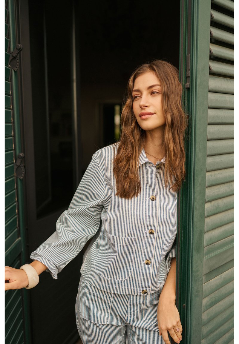 Junge Frau mit welligem Haar, die eine gestreifte, geknöpfte Jacke und Hose trägt, steht in einer teilweise geöffneten grünen Tür mit Fensterläden.