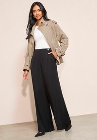 Kostkovaná hnědá bunda s detailem knoflíků, spárovaná s širokými černými kalhotami. Modelka má na sobě bílé tričko a černé špičaté lodičky. Jednoduché pozadí.