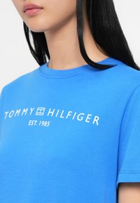 Młoda kobieta w jasnoniebieskiej koszulce Tommy Hilfiger z białym logo i napisem "EST. 1985", widoczna od ramion do warg.