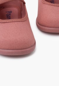 Zapatillas de lona rosas con una textura suave y una puntera redondeada. Cuentan con una correa en la parte superior y una suela de goma. Detalles de costura notables.