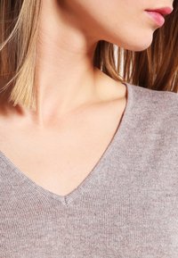 Jeune femme aux cheveux blonds raides portant un pull en tricot marron clair à col en V, tête tournée sur le côté montrant la ligne de la mâchoire et du cou.