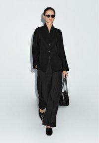 Blazer nero oversize con rever, chiusura con un bottone, pantaloni neri larghi, scarpe nere e una piccola borsa a tracolla. Occhiali da sole indossati.