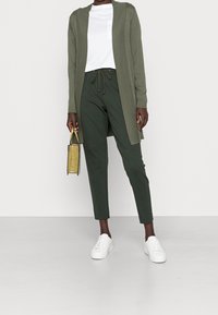 Olivengrøn cardigan over en hvid t-shirt, kombineret med mørkegrønne bukser og hvide sneakers. Holder en lille gul håndtaske.