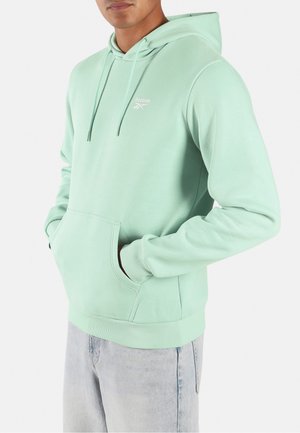 Hombre con sudadera con capucha Reebok verde claro con bolsillo frontal y cordones, combinado con vaqueros de mezclilla azul claro.