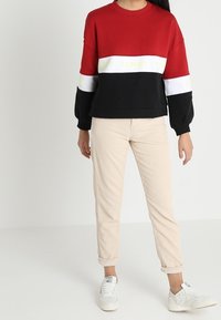 Felpa a blocchi di colore rossa e nera con striscia bianca e testo giallo. Tessuto leggero e lavorato a maglia, abbinata a pantaloni beige e sneakers bianche.