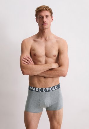 Bovenlijfs man met een lichte huid en kort krullend haar, staand met gekruiste armen in grijze MARC O’POLO boxershorts tegen een eenvoudige witte achtergrond.