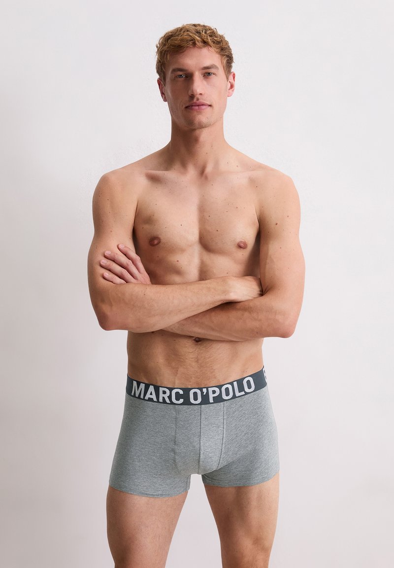 Bovenlijfs man met een lichte huid en kort krullend haar, staand met gekruiste armen in grijze MARC O’POLO boxershorts tegen een eenvoudige witte achtergrond.
