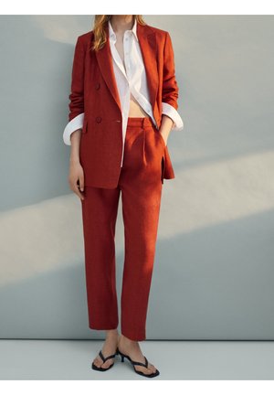 Kobieta w rdzawej marynarce z podwiniętymi rękawami, białej częściowo rozpiętej koszuli i czarnych sandałkach na obcasie, stojąca przy szarej ścianie.