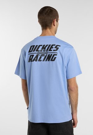 Man som bär en ljusblå t-shirt med svart text ""Dickies Racing"" och rutigt logotyp på baksidan, kombinerad med svarta byxor.