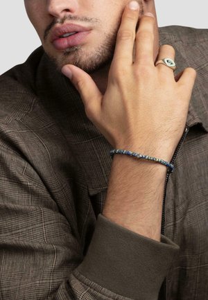 Beaded bracelet with blue and green tones, worn on a hand with light skin. The fabric is a gray plaid shirt with a dark collar.