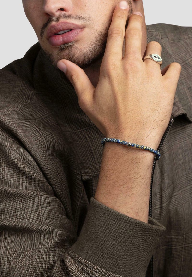 Beaded bracelet with blue and green tones, worn on a hand with light skin. The fabric is a gray plaid shirt with a dark collar.