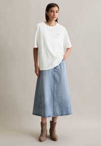 T-shirt en coton blanc à manches courtes avec une poche, associé à une longue jupe en denim bleu clair et des bottines beiges à la cheville.