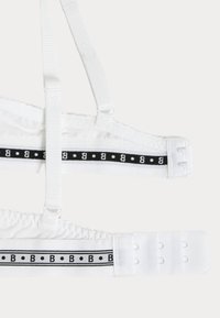 Bralette blanche avec des bretelles réglables et une bande élastique à motifs noir et blanc présentant de petits points et des accents de lettres sur la bande.