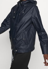 Marinblå vattentät jacka gjord av slätt material, med huva, framfickor, knäppning med knappar och justerbar dragsko i nederkanten.