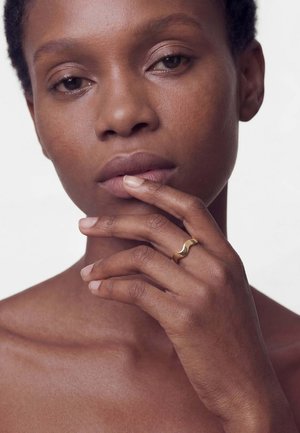 Mujer con tono de piel natural tocándose los labios, con un anillo dorado ondulado en el dedo, sobre un fondo blanco liso.