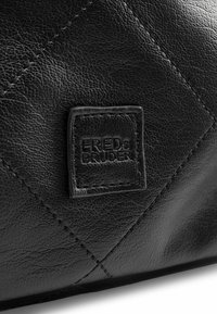 Bolso de cuero negro con un patrón de diamantes y una superficie texturizada. Incluye una etiqueta cuadrada con relieve que dice "FREDs BRUDER".