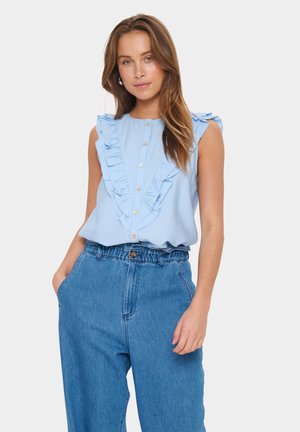 Ljusblå ärmlös blus med volangdetaljer, med en knäppt front och en avslappnad passform, som är parat med högsadlade jeansbyxor.
