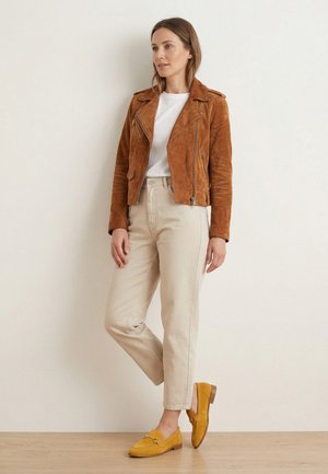 Kvinde iført en brun ruskindjakke, hvid skjorte, beige bukser og sennepsgule loafers, står op ad en almindelig væg med det ene ben krydset.