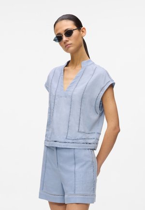 Svetlo modra dvodelna lanena obleka s kratkimi rokavi, V-izrezom in ujemajočimi kratkimi hlačami. Poudarjena z dekorativnim šivom.