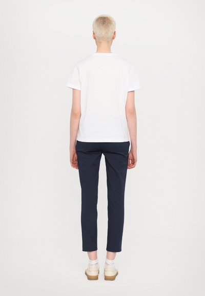 Hvid bomulds T-shirt med korte ærmer, parret med navy bukser. Modellen har beige fodtøj på med hvide sokker, vist fra ryggen.