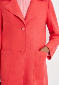 Coral wool coat with a notched collar, two buttons, and patch pockets. Features a soft texture and structured design.