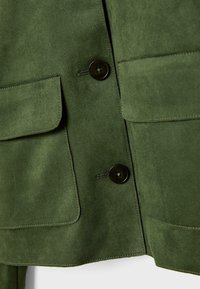 Veste en daim vert olive avec deux poches avant, boutons noirs, et une texture lisse avec des détails de surpiqûres visibles.