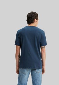 Hombre con cabello oscuro que lleva una camiseta azul marino sencilla y jeans de color azul claro, de espaldas sobre un fondo gris neutro.