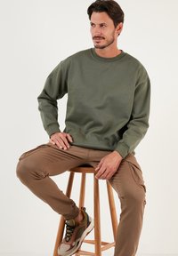 Sudadera verde oliva con cuello redondo y puños, combinada con pantalones cargo marrones y zapatillas multicolores. Fondo de un taburete de madera elegante.