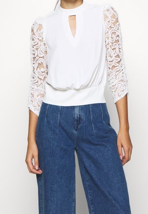 Femme portant un haut blanc avec manches 3/4 en dentelle et un décolleté en forme de trou de serrure, associé à un pantalon large en jean bleu taille haute.