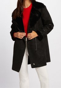 Sort faux shearling frakke med bred krave og lynlås lukning. Har en glat tekstur og pelsfor, parret med en rød ribbet sweater.