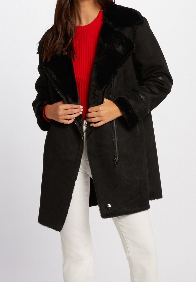 Sort faux shearling frakke med bred krave og lynlås lukning. Har en glat tekstur og pelsfor, parret med en rød ribbet sweater.