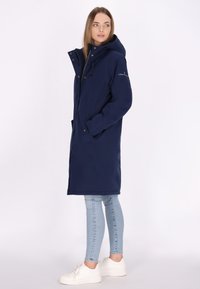 Marineblauwe lange jas met capuchon, voorzien van een ritssluiting en drukknoopsluiting, zijzakken en een soepele textuur. Gecombineerd met lichtblauwe jeans en witte sneakers.