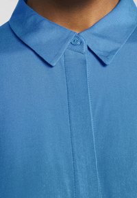 Chemise bleu col avec un tissu lisse. Elle comporte une fermeture à boutons et un col structuré avec une légère brillance et une texture nette.