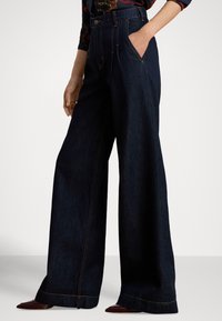 Høytlivs, vide jeans i mørk denim med lys oransje søm og sidelommer. Jevn tekstur og rett snitt fra midje til kant.