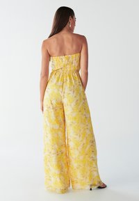 Geltonas, be petnešėlių kombinezonas su surištu korpusu ir plačiomis kelnėmis, puoštas gėlių raštu šviesiomis ir tamsiomis spalvų atspalviais.