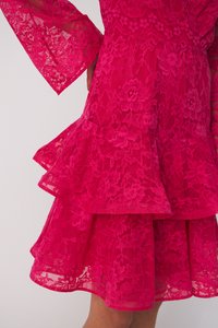 Rochie din dantelă roz aprins cu volane suprapuse la tiv și mâneci clopot, având modele florale și o textură finisată.