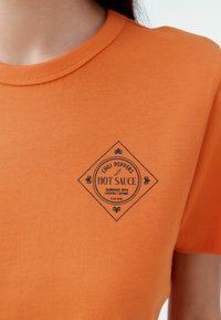 Oranssi puuvillapaita, jossa on musta timantinmuotoinen grafiikka, jossa lukee "Hot Sauce" ja koristeelliset yksityiskohdat. Pehmeä tekstuuri, klassinen pyöreä kaula-aukko.